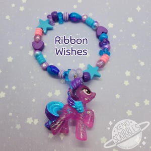May include: A purple and blue pony charm hangs from a colorful beaded bracelet. The bracelet features blue, purple, pink, and white beads in various shapes, including stars and hearts. The text "Ribbon Wishes" is printed on the bracelet.