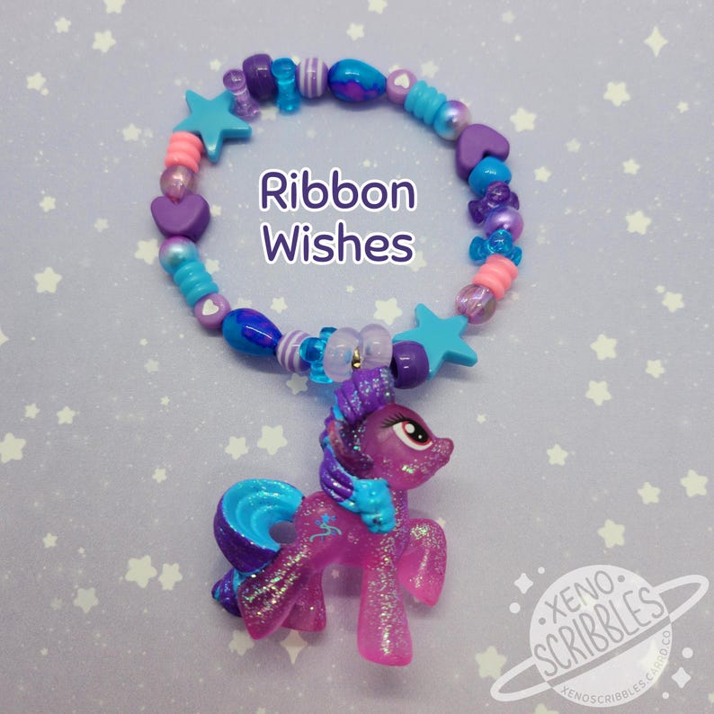 May include: A purple and blue pony charm hangs from a colorful beaded bracelet. The bracelet features blue, purple, pink, and white beads in various shapes, including stars and hearts. The text "Ribbon Wishes" is printed on the bracelet.