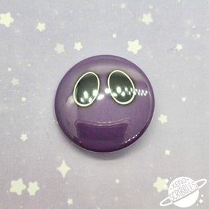 May include: Purple button with a cartoon face with two large black eyes. The button is on a purple background with white stars.