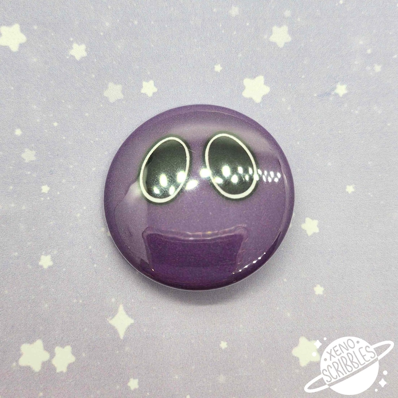May include: Purple button with a cartoon face with two large black eyes. The button is on a purple background with white stars.