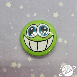 May include: Green button with a smiling face with large blue eyes and a wide white smile. The button has a glossy finish and is on a purple background with white stars.