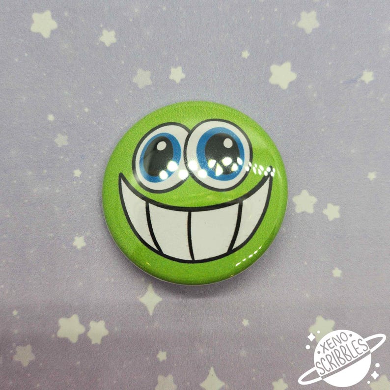May include: Green button with a smiling face with large blue eyes and a wide white smile. The button has a glossy finish and is on a purple background with white stars.