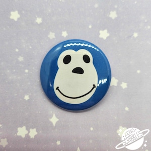 May include: Blue button with a white cartoon monkey face with black eyes and a black nose. The monkey is smiling.