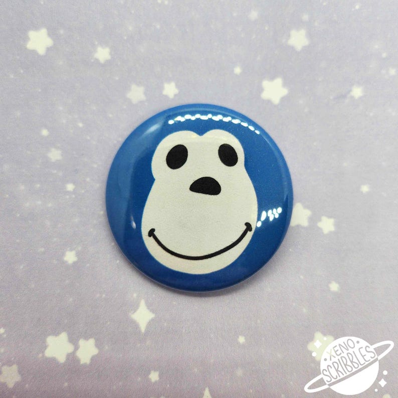 May include: Blue button with a white cartoon monkey face with black eyes and a black nose. The monkey is smiling.
