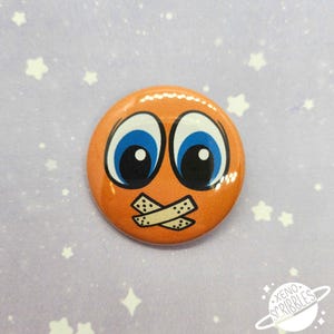 May include: Orange button with a cartoon face with blue eyes and a piece of tape over the mouth.