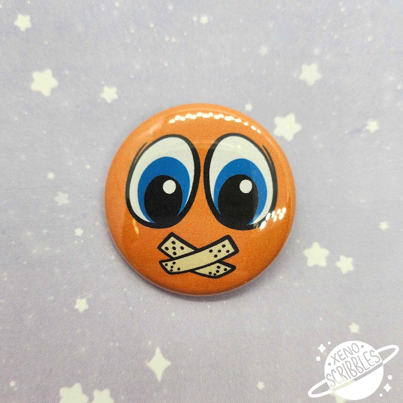 May include: Orange button with a cartoon face with blue eyes and a piece of tape over the mouth.