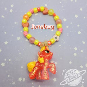 May include: A pink and yellow beaded bracelet with a pink heart charm and a small pink and yellow pony charm. The bracelet is labeled "Junebug".