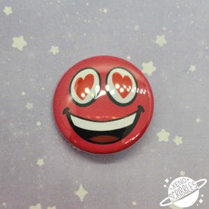 May include: Red smiley face button with heart eyes. The button is round and has a white background with a red smiley face with white teeth and red heart eyes.