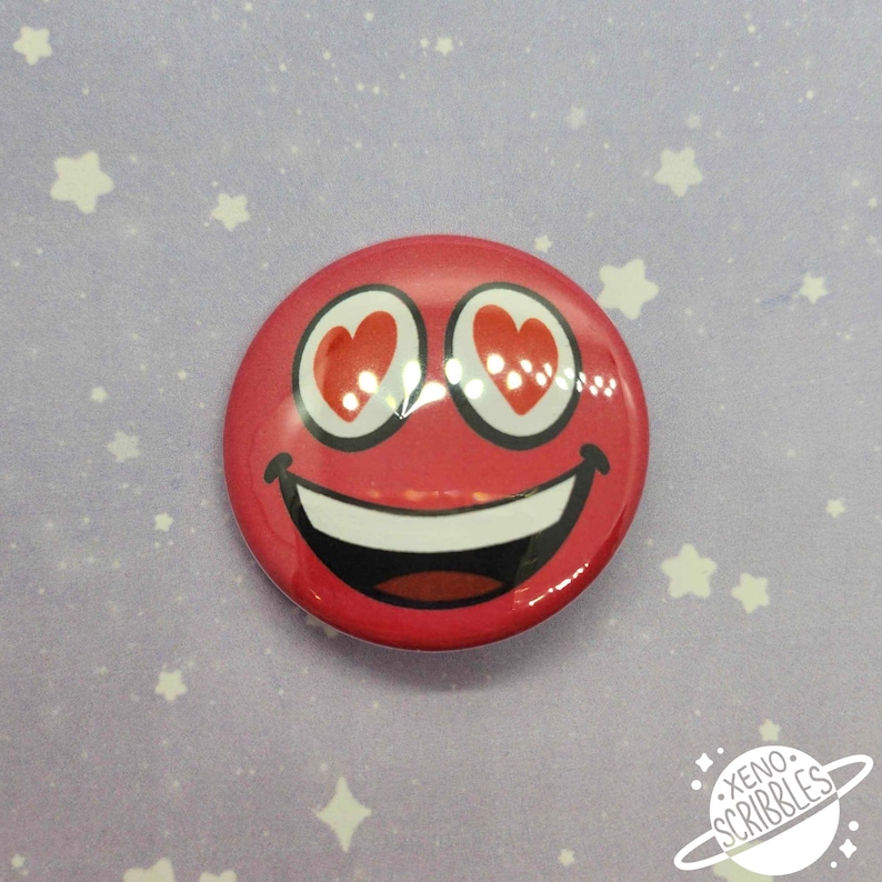 May include: Red smiley face button with heart eyes. The button is round and has a white background with a red smiley face with white teeth and red heart eyes.