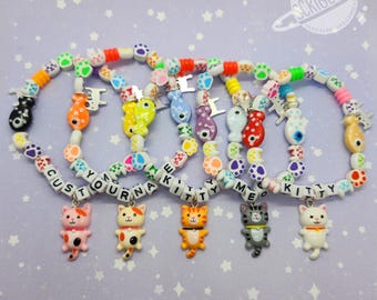 May include: Six colorful beaded bracelets with cat charms. Each bracelet features a different color scheme, with paw print beads and letter beads spelling out "CUSTOM", "YOUR", "KITTY", and "MEOW KITTY". The charms are various cat designs.