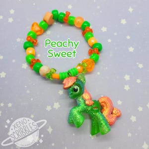 May include: A green and orange beaded bracelet with a small green glittery pony charm. The bracelet has the text "Peachy Sweet" on it.
