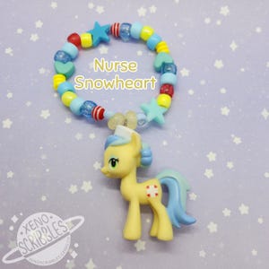 May include: A yellow pony with blue hair and a nurse's hat is attached to a colorful beaded bracelet. The bracelet has red, yellow, blue, and green beads, as well as stars and hearts. The text "Nurse Snowheart" is written on the bracelet.
