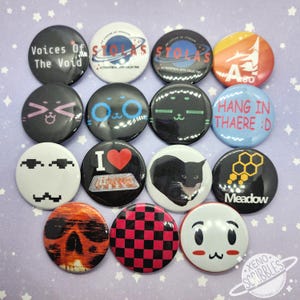 Puede incluir: Una colección de botones redondos con varios diseños. Algunos botones presentan texto como "Voices of The Void" y "HANG IN THAERE :D". Otros diseños incluyen un gato, una calavera y un patrón de cuadros rojos y negros. Los botones son de varios colores.