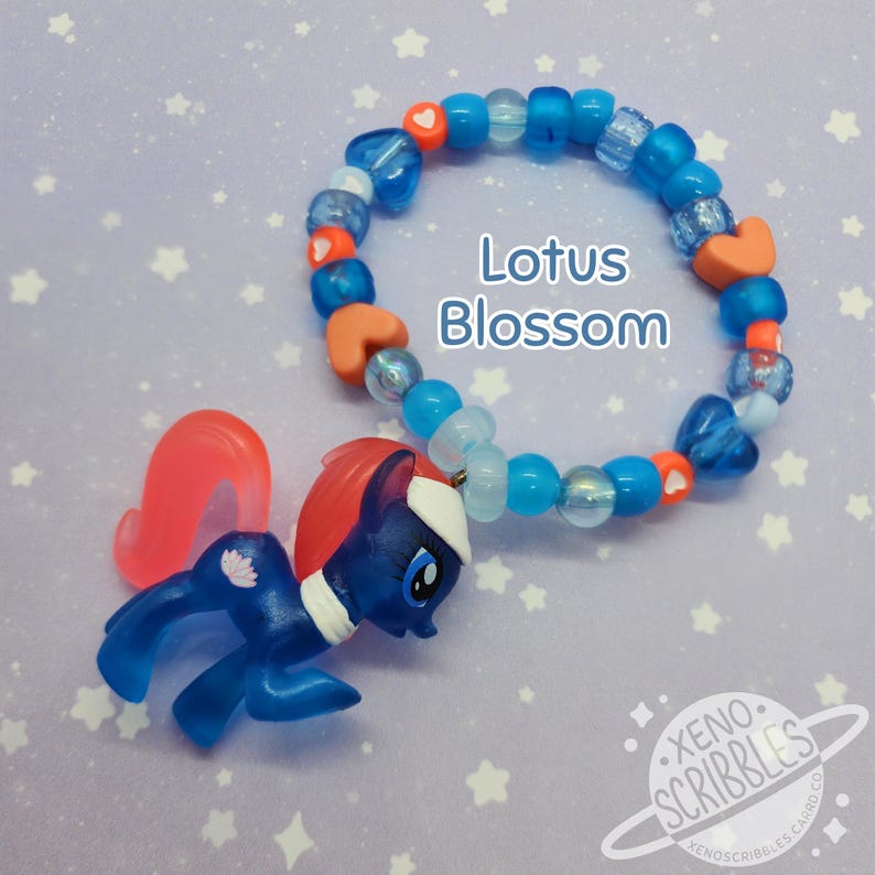 May include: A blue and orange beaded bracelet with a charm of a blue pony with a pink mane and tail. The bracelet has white hearts and blue beads. The charm has a lotus flower on its side. The text "Lotus Blossom" is written on the image.