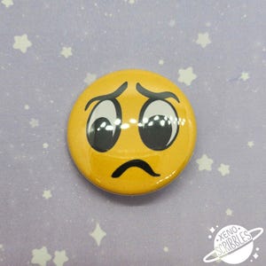 May include: Yellow button with a cartoon face expressing sadness. The face has large, wide eyes and a downturned mouth.