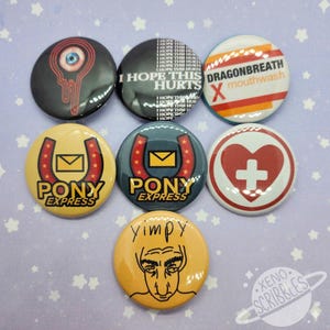May include: Set of 7 colourful button badges with various designs. The badges feature a red eye with dripping liquid, a yellow horseshoe with a red star and a red envelope, a blue horseshoe with a red star and a red envelope, a red heart with a white cross, a yellow face with a black outline, a black and white badge with the text "I hope this hurts" and a red, orange and white badge with the text "Dragonbreath X mouthwash".