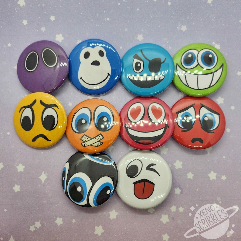 May include: Set of ten 1-inch round button pins featuring various colorful cartoon faces with different expressions. The faces include a monkey, a pirate, a winking face, a sad face, a face with hearts for eyes, a face with tape over its mouth, a face with googly eyes, a purple face with two dots for eyes, a red face with tears, and a white face with a tongue sticking out.