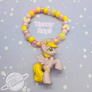 May include: A pink and yellow beaded bracelet with a heart charm and a small pink pony figurine with yellow hair. The bracelet has the text "Honey Rays" on it.
