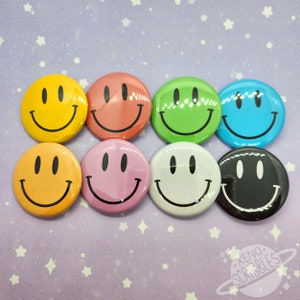 Könnte beinhalten: Acht runde Ansteckbuttons mit Smiley-Gesichtern in verschiedenen Farben: Gelb, Orange, Rosa, Rot, Grün, Blau, Weiß und Schwarz. Jeder Button hat ein schwarzes Smiley-Gesicht auf einem einfarbigen Hintergrund. Die Buttons sind in zwei Reihen zu je vier Stück angeordnet.