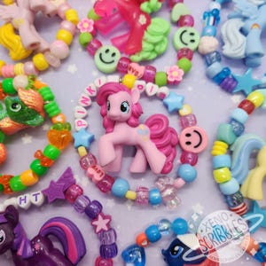 May include: A collection of colorful beaded bracelets with charms, including a pink pony, a smiley face, and stars. The bracelets are made with various colors of beads, including pink, blue, green, and yellow.