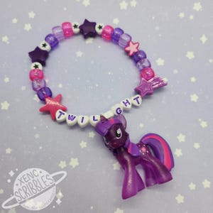 May include: A purple and pink beaded bracelet with a star charm and a small purple Twilight Sparkle pony figure. The bracelet spells out "TWILIGHT" in white letters.