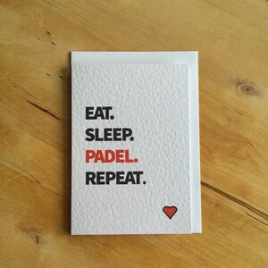 May include: A white greeting card with the words "EAT. SLEEP. PADEL. REPEAT." in black text, with the word "PADEL" in red. A small red heart is at the bottom. The card is on a wooden surface.