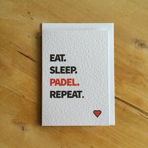 May include: A white greeting card with the words "EAT. SLEEP. PADEL. REPEAT." in black text, with "PADEL" in red. A small red heart is at the bottom. The card is on a wooden surface.