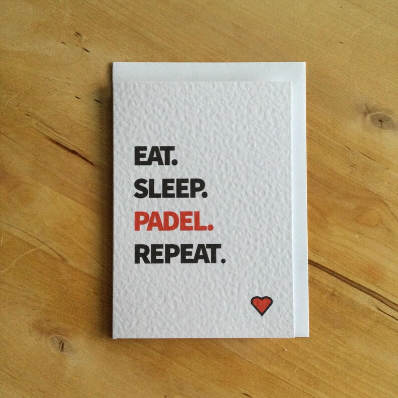 May include: A white greeting card with the words "EAT. SLEEP. PADEL. REPEAT." in black text, with "PADEL" in red. A small red heart is at the bottom. The card is on a wooden surface.