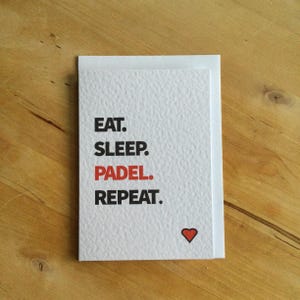 May include: A white card with the text "EAT. SLEEP. PADEL. REPEAT." in black, with "PADEL" in red. A small red heart is at the bottom. The card is on a wooden surface.