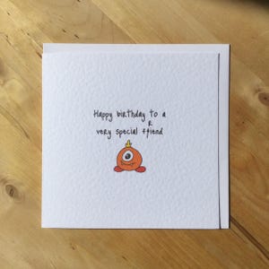 May include: A white greeting card with the handwritten message "Happy birthday to a very special friend." The card features a small, orange cartoon monster with one eye and a small crown. The card is on a wooden surface.