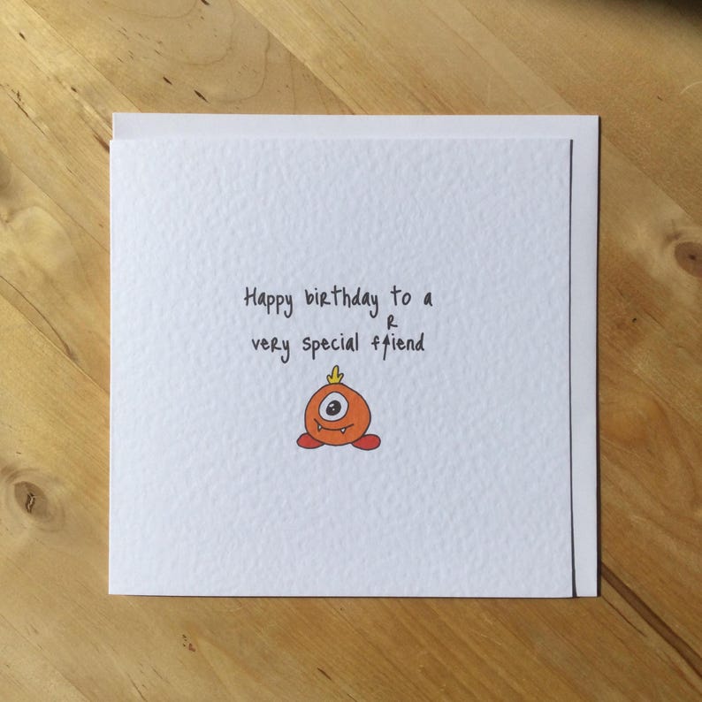 May include: A white greeting card with the handwritten message "Happy birthday to a very special friend." The card features a small, orange cartoon monster with one eye and a small crown. The card is on a wooden surface.