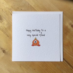 May include: A white greeting card with the text "Happy birthday to a very special friend." The card features a cartoon orange monster with one eye and a small crown. The card is on a wooden surface.