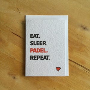 May include: White greeting card with the words "EAT. SLEEP. PADEL. REPEAT." in black and red text. A small red heart is in the lower right corner. The card is on a wooden surface.
