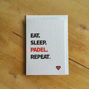 May include: A white card with the text "EAT. SLEEP. PADEL. REPEAT." in black and red. A small red heart is in the lower right corner. The card is on a wooden surface.