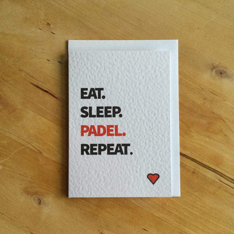 May include: A white card with the text "EAT. SLEEP. PADEL. REPEAT." in black and red. A small red heart is in the lower right corner. The card is on a wooden surface.