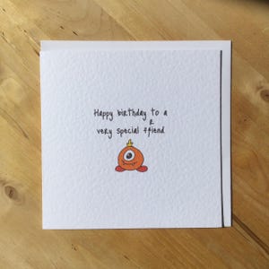 May include: A white greeting card with the text "Happy birthday to a very special friend." A small, orange monster illustration with one eye and red feet is below the text. The card is on a wooden surface.