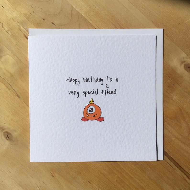 May include: A white greeting card with the text "Happy birthday to a very special friend." A small, orange monster illustration with one eye and red feet is below the text. The card is on a wooden surface.