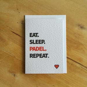 May include: A white card with the text "EAT. SLEEP. PADEL. REPEAT." in black and red. A small red heart is in the lower right corner. The card is on a wooden surface.