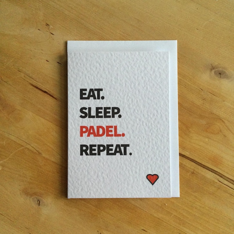 May include: A white card with the text "EAT. SLEEP. PADEL. REPEAT." in black and red. A small red heart is in the lower right corner. The card is on a wooden surface.