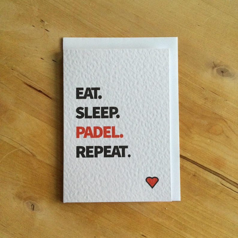 May include: White greeting card with the words "EAT. SLEEP. PADEL. REPEAT." in black text, with "PADEL" in red. A small red heart is at the bottom right. The card is on a wooden surface.