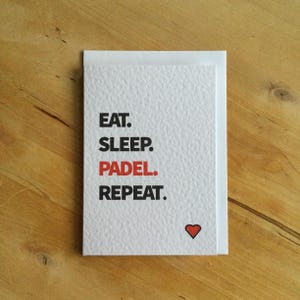 May include: A white card with the text "EAT. SLEEP. PADEL. REPEAT." in black and red. A small red heart is at the bottom right. The card is on a wooden surface.