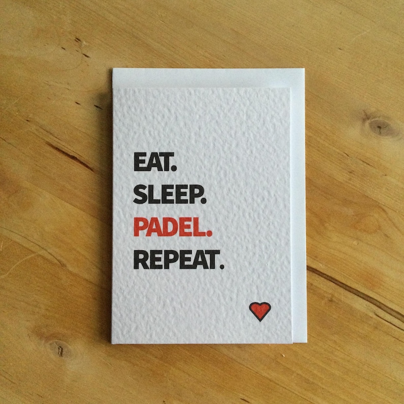 May include: A white card with the text "EAT. SLEEP. PADEL. REPEAT." in black and red. A small red heart is at the bottom right. The card is on a wooden surface.