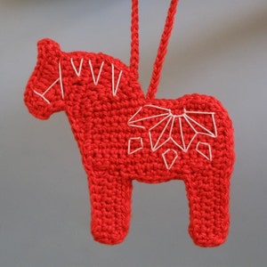 May include: Red crocheted horse ornament with white embroidery. The horse is a traditional Swedish Dala horse, a popular folk art motif.