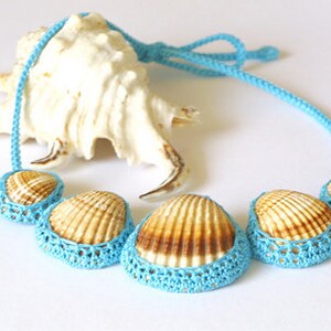 May include: A blue crocheted necklace with five seashells. The shells are brown and have a natural texture. The necklace is on a white background.