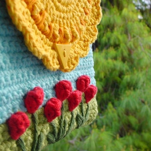 May include: A crocheted bag with a blue and yellow floral design. The bag has a yellow crocheted flap with a plastic clasp. The bag is decorated with red crocheted flowers.