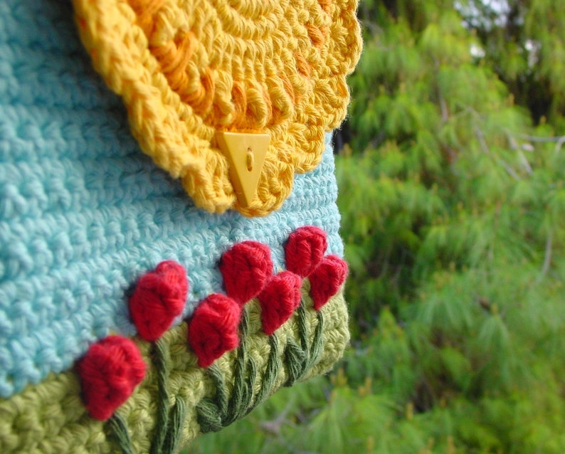 May include: A crocheted bag with a blue and yellow floral design. The bag has a yellow crocheted flap with a plastic clasp. The bag is decorated with red crocheted flowers.