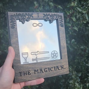 May include: A wooden mirror with a dark brown stain. The mirror has a carved frame with a leafy design and the text "I ∞ THE MAGICIAN." The mirror also has a pentagram, two swords, and a goblet carved into the surface.