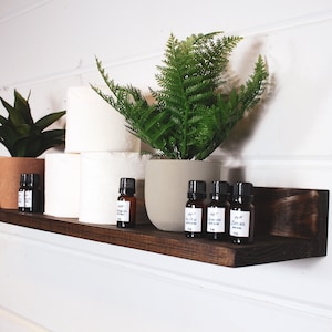 May include: A dark brown wooden shelf mounted on a white wall with a variety of items on it, including two potted plants, a stack of toilet paper rolls, and several small bottles of essential oils.