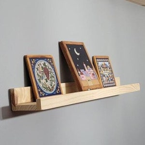 May include: A wooden shelf with three framed pictures. The shelf is light brown and has a slightly curved edge. The pictures are all different sizes and have different designs. The shelf is mounted on a gray wall.