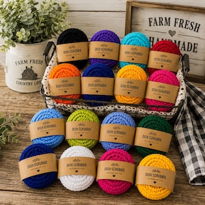 May include: A collection of round, crocheted dish scrubbies in various solid colors, including black, purple, turquoise, red, orange, blue, yellow, pink, light blue, lime green, dark blue, white, and dark green, each wrapped in a brown paper label.